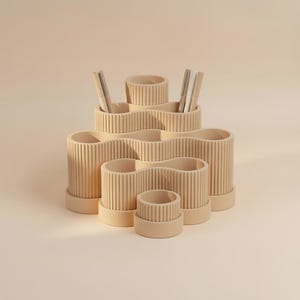 Pode incluir: Um organizador de secretária bege com um design em camadas de vários compartimentos e texturas caneladas verticais. A estrutura consiste em vários suportes cilíndricos interligados de alturas variadas, com duas canetas na parte superior.