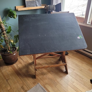 Peut inclure: Une table à dessin vintage avec une surface noire et un cadre en bois. La table est inclinée, avec un petit motif floral sur le dessus. Elle repose sur un plancher en bois.