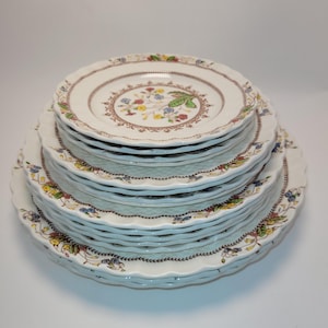 May include: A collection of white ceramic plates with a floral pattern. The plates have a scalloped edge and a brown border with a repeating design. The center of the plates features a colorful floral arrangement.