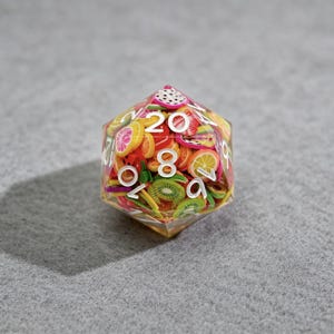 Op de afbeelding: Een heldere, veelzijdige dobbelsteen gevuld met kleurrijke fruitschijfjes, waaronder kiwi, citroen en drakenfruit. De dobbelsteen heeft witte cijfers en staat tegen een grijze achtergrond. Een uniek tabletop gaming accessoire.