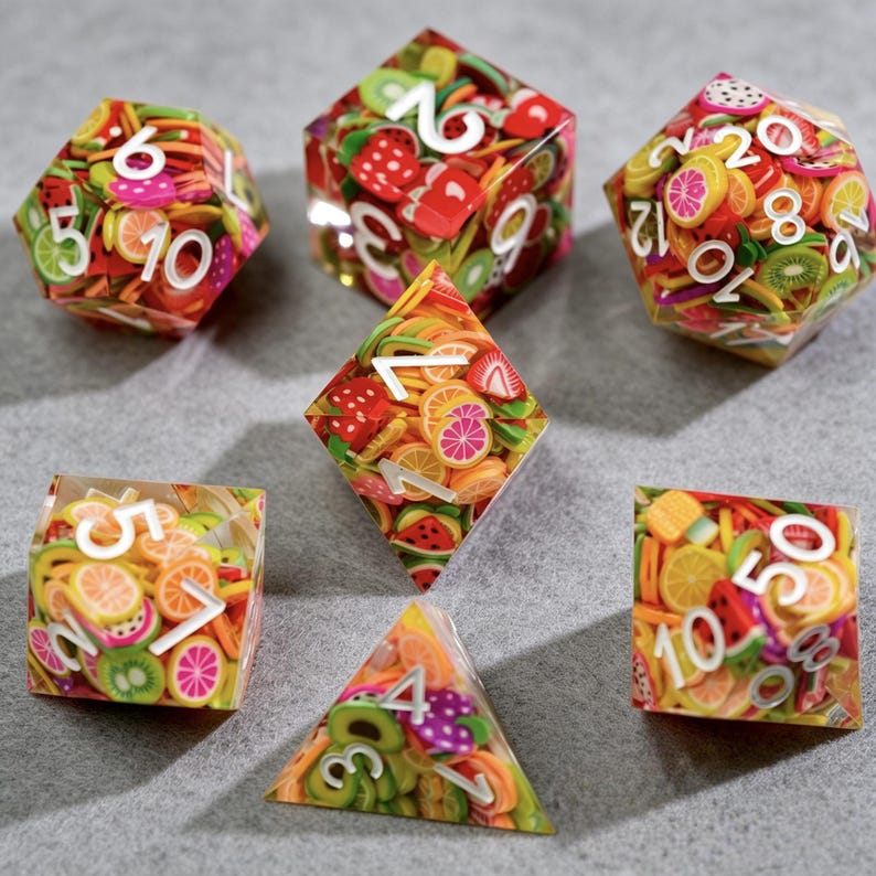 Op de afbeelding: Een verzameling kleurrijke dobbelstenen met fruitplakontwerpen. De dobbelstenen zijn transparant met verschillende fruitvormen en witte cijfers. De dobbelstenen hebben verschillende geometrische vormen, waaronder een d20, d12, d10, d8, d6 en een piramidevormige d4.
