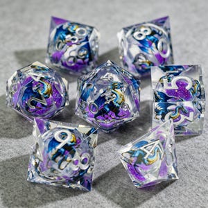 Puede incluir: Juego de siete dados poliédricos con temática de fantasía. Cada dado es transparente con un diseño de dragón y galaxia en azul, morado y plateado. Los números son plateados. Los dados son para juegos de rol de mesa.
