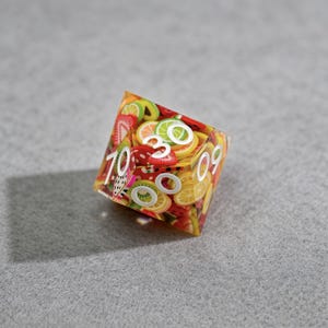 Op de afbeelding: Een veelzijdige dobbelsteen met een transparant ontwerp met fruit als thema. De dobbelsteen is bedekt met plakjes verschillende soorten fruit, waaronder aardbeien, citroenen en kiwi's. Witte cijfers zijn zichtbaar op de zijkanten. De dobbelsteen staat tegen een grijze achtergrond.