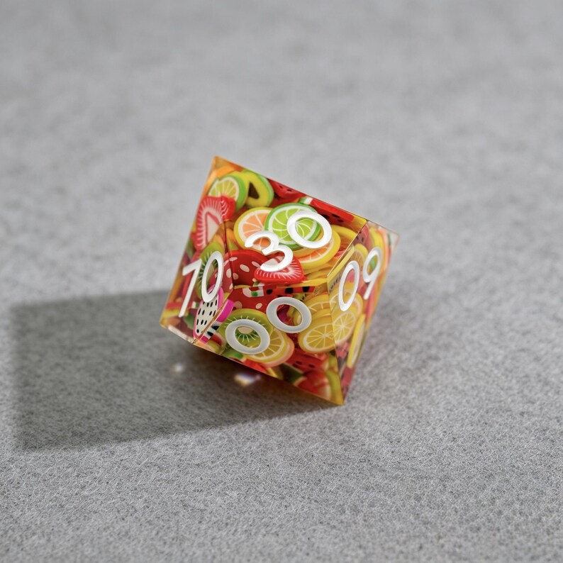 Op de afbeelding: Een veelzijdige dobbelsteen met een transparant ontwerp met fruit als thema. De dobbelsteen is bedekt met plakjes verschillende soorten fruit, waaronder aardbeien, citroenen en kiwi's. Witte cijfers zijn zichtbaar op de zijkanten. De dobbelsteen staat tegen een grijze achtergrond.