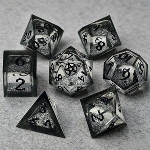 Può includere: Un set di sette dadi poliedrici realizzati in resina trasparente con bordi neri e riempimento di glitter argento. I dadi presentano numeri neri e il dado a venti facce mostra un simbolo di pentagramma nero sulla sua faccia superiore.