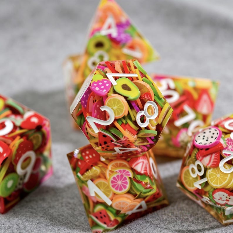 Op de afbeelding: Een set kleurrijke dobbelstenen met fruit thema. Elke dobbelsteen is transparant en bevat miniatuur fruitplakjes, waaronder aardbeien, watermeloen en avocado. Witte cijfers zijn zichtbaar op elke zijde. De dobbelstenen liggen op een grijze ondergrond.