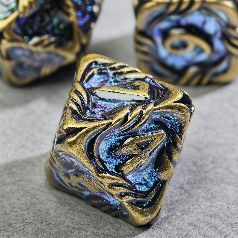May include: Close-up of several square-shaped beads with a swirling, textured design. The beads feature a metallic gold finish with iridescent blue and purple accents, creating a unique, eye-catching appearance. These beads are ideal for jewelry making.