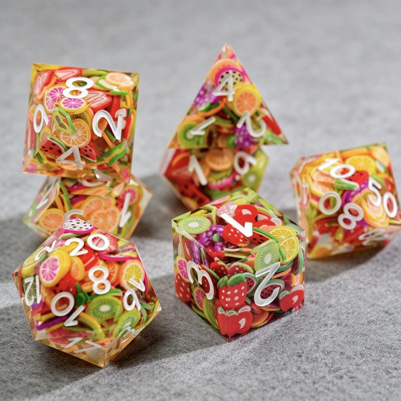 Op de afbeelding: Een verzameling polyhedrale dobbelstenen met een fruitig ontwerp. De transparante dobbelstenen zijn gevuld met kleurrijke fruitschijfjes, waaronder aardbeien, kiwi's en sinaasappels. Witte cijfers zijn zichtbaar op elke zijde. De dobbelstenen liggen op een grijze ondergrond.