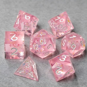 Peut inclure: Un ensemble de sept dés polyédriques en résine rose translucide. Chaque dé contient des confettis colorés et des paillettes en forme d'étoile. Les chiffres sont gravés dans une police blanche nette, offrant un contraste élevé avec le matériau rose.