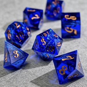 Könnte beinhalten: Set aus sieben blauen, polyedrischen Würfeln mit goldfarbenen Zahlen. Die Würfel sind transparent und haben ein wirbelndes, strukturiertes Inneres. Die Würfel liegen auf einer grauen Oberfläche.