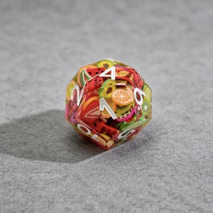 Op de afbeelding: Een veelzijdige dobbelsteen met een transparante buitenkant, gevuld met kleurrijke fruitschijfjes. De dobbelsteen heeft witte cijfers en een verscheidenheid aan fruit, waaronder watermeloen, sinaasappels en kiwi. De dobbelsteen staat tegen een grijze achtergrond.
