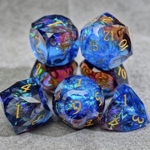 Puede incluir: Juego de siete dados poliédricos con un esquema de color azul e iridiscente. Los dados tienen numeración dorada y un diseño facetado. El juego incluye un D4, D6, D8, D10, D12, D20 y un dado de porcentaje.