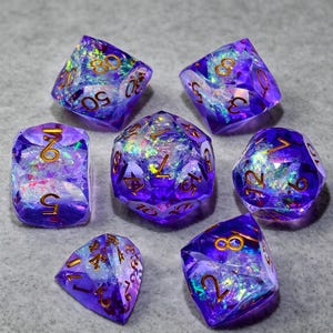 May include: A set of seven polyhedral dice in a translucent purple resin with embedded iridescent foil flakes that catch the light. Each die features sharp edges and clear, gold-colored numbers engraved on the faces for easy readability.