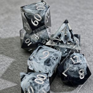 May include: A set of seven polyhedral dice for tabletop games. The dice are black with a swirling white pattern and silver numbering. The dice include a d4, d6, d8, d10, d12, d20, and a percentile die.
