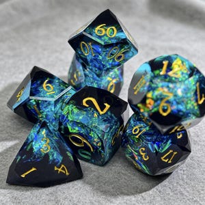 Könnte beinhalten: Ein Satz von sieben polyedrischen Würfeln mit einem dunkelblauen und grünen schillernden Design. Die Würfel haben goldene Zahlen und sind für Tabletop-Rollenspiele konzipiert. Die Würfel werden auf einer grauen Oberfläche gezeigt.