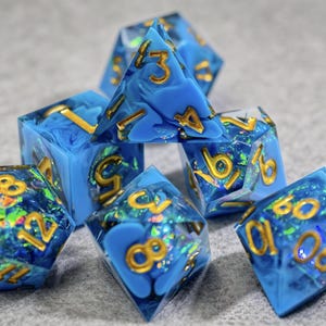 May include: Set of seven polyhedral dice with a blue and white marbled design, gold numbering, and iridescent glitter inclusions. The dice are for tabletop games.
