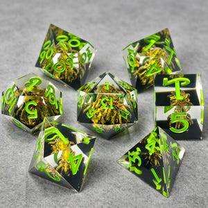 May include: A set of seven polyhedral dice made of clear resin, each containing a miniature golden spider figure with red eyes. The numbers on the dice are engraved and filled with a bright neon green pigment, providing high contrast.