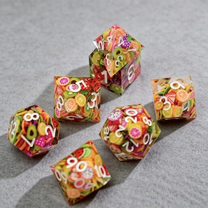 Op de afbeelding: Een set van zeven polyhedrale dobbelstenen met een fruitsalade thema. Elke dobbelsteen is transparant met kleurrijke fruitschijfjes en witte cijfers. Dobbelstenen zijn onder andere een D4, D6, D8, D10, D12, D20 en een D6 kubus.