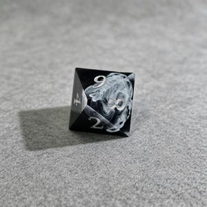 May include: A black and white D20 dice with silver numbering. The die features a swirling white pattern on one face, with the number 8 prominently displayed. The dice is designed for tabletop games.