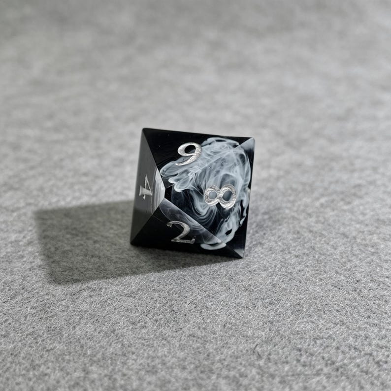 May include: A black and white D20 dice with silver numbering. The die features a swirling white pattern on one face, with the number 8 prominently displayed. The dice is designed for tabletop games.