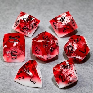 Puede incluir: Un juego de siete dados poliédricos con un degradado de blanco opaco en la base a rojo translúcido en la parte superior, con copos de papel de plata incrustados y números negros grabados. El set incluye un d20, d12, d10, d8, d6, d4 y d100.