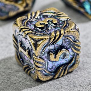 May include: Close-up of a square, ornate die with a metallic gold and iridescent finish. The die features intricate floral and scrollwork designs on each face, with numbers in gold. The overall color scheme is a mix of gold, purple, and blue.