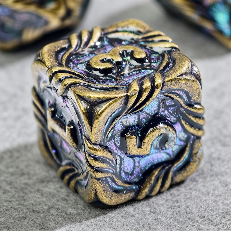 May include: Close-up of a square, ornate die with a metallic gold and iridescent finish. The die features intricate floral and scrollwork designs on each face, with numbers in gold. The overall color scheme is a mix of gold, purple, and blue.