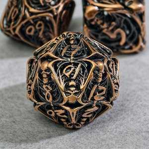Puede incluir: Primer plano de un dado poliédrico de bronce y negro con diseños de calaveras y adornos. El dado presenta números y símbolos, con un acabado metálico. Los dados se utilizan probablemente para juegos de mesa.