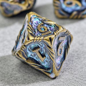 May include: Close-up of a polyhedral die with a metallic gold and iridescent finish. The die features the number 6 on multiple faces. The surface has a textured, carved design. Other dice are visible in the background.