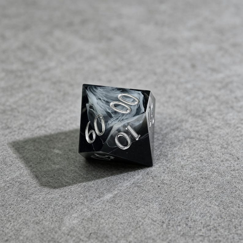 May include: A black and white marbled D6 dice with silver numbering. The dice has the numbers 0, 10, and 60 visible. The dice is sitting on a gray surface.