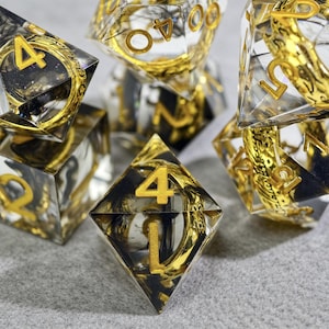 May include: A set of clear resin dice with gold-colored numbers and intricate gold-colored designs inside. The dice have a black and gold color scheme, with the numbers prominently displayed. The dice are designed for tabletop gaming.
