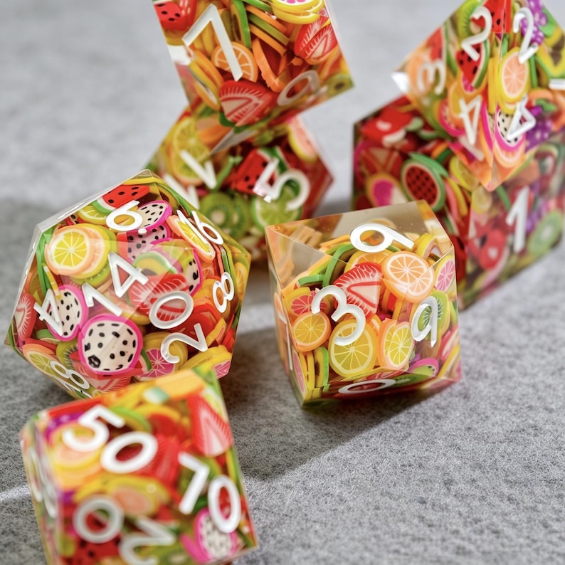 Op de afbeelding: Een verzameling dobbelstenen met een ontwerp van fruitschijfjes. De transparante dobbelstenen zijn gevuld met kleurrijke fruitschijfjes en witte cijfers. Vruchten zijn onder andere watermeloen, kiwi en sinaasappels. Voor tabletop games.