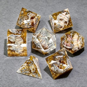 Könnte beinhalten: Ein Set aus sieben polyedrischen Würfeln aus klarem Harz, die jeweils kleine Muscheln und goldene Sandpartikel enthalten. Die Würfel haben silberfarbene, in die Oberflächen eingravierte Zahlen, darunter ein W20, W12, W10, W8, W6, W4 und ein Prozentwürfel.