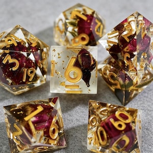 May include: A set of seven polyhedral dice. Each die is clear with gold numbering and gold flakes, and contains dried red rose petals. The dice are in various geometric shapes, including a cube, a d20, and a d12.