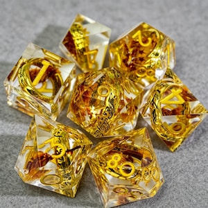 May include: Set of seven clear resin dice with gold-colored numbers and decorative elements. The dice have a mix of gold and brown inclusions, with a gold-colored font. The dice are designed for tabletop gaming.