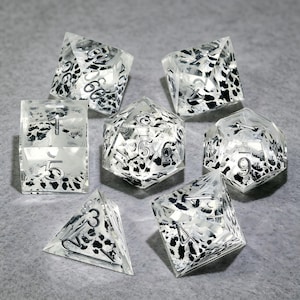 Könnte beinhalten: Ein Set aus sieben polyedrischen Würfeln aus klarem Harz mit schwarzen und weißen gesprenkelten Einschlüssen. Jeder Würfel hat scharfe Kanten und eingravierte silberfarbene Zahlen auf den Flächen, darunter W20, W12, W10, W8, W6, W4 und ein Prozentwürfel.