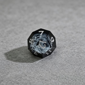 May include: A black and white twelve-sided die with silver numbering. The die has a smoky white interior and is resting on a gray surface. The numbers are clearly visible, making it suitable for tabletop games.