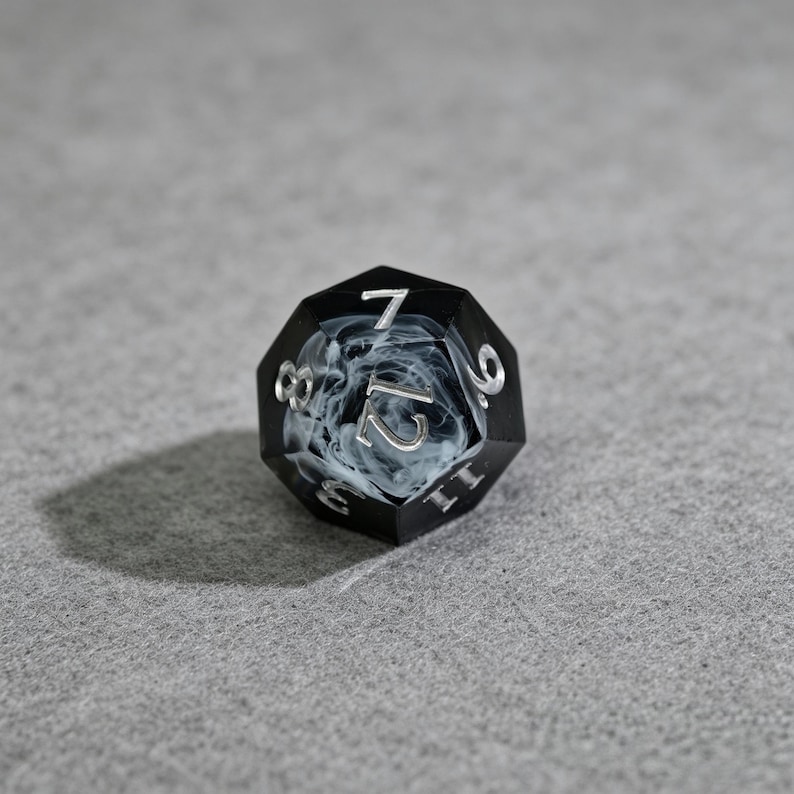 May include: A black and white twelve-sided die with silver numbering. The die has a smoky white interior and is resting on a gray surface. The numbers are clearly visible, making it suitable for tabletop games.