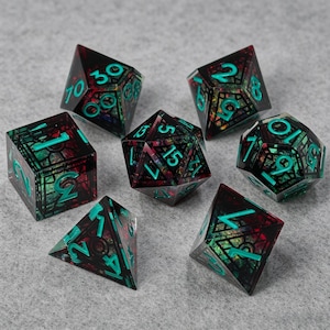 Pode incluir: Um conjunto de sete dados poliédricos com uma base de resina escura e translúcida, flocos de folha iridescentes e multicoloridos incrustados e números em um tom brilhante de verde-azulado. O conjunto inclui um d20, d12, d10, d8, d6, d4 e um dado percentual.