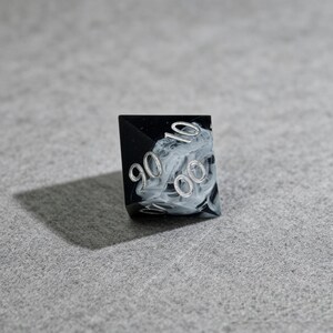 May include: A black and white marbled polyhedral die with silver numbering. The die features the numbers 90, 10, and 00. The die is set against a gray background.