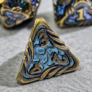 May include: Close-up of a bronze-colored, triangular die with blue and gold accents. The die features the number 3 in a stylized font. The edges are adorned with intricate patterns, and the die has a metallic sheen. Other dice are visible in the background.