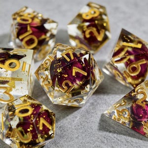 Può includere: Set di sette dadi poliedrici. Ogni dado è trasparente con numeri dorati e contiene un fiore rosso essiccato e scaglie d'oro. I dadi hanno varie forme geometriche, tra cui un d20, d10, d8, d6 e d4.