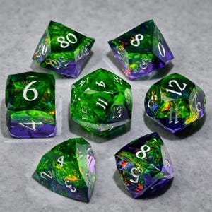May include: A set of seven polyhedral dice featuring a translucent green and purple color gradient with embedded iridescent foil flakes. Each die has sharp edges and clear, white engraved numbers, including a d20, d12, d10, d8, d6, d4, and d100.