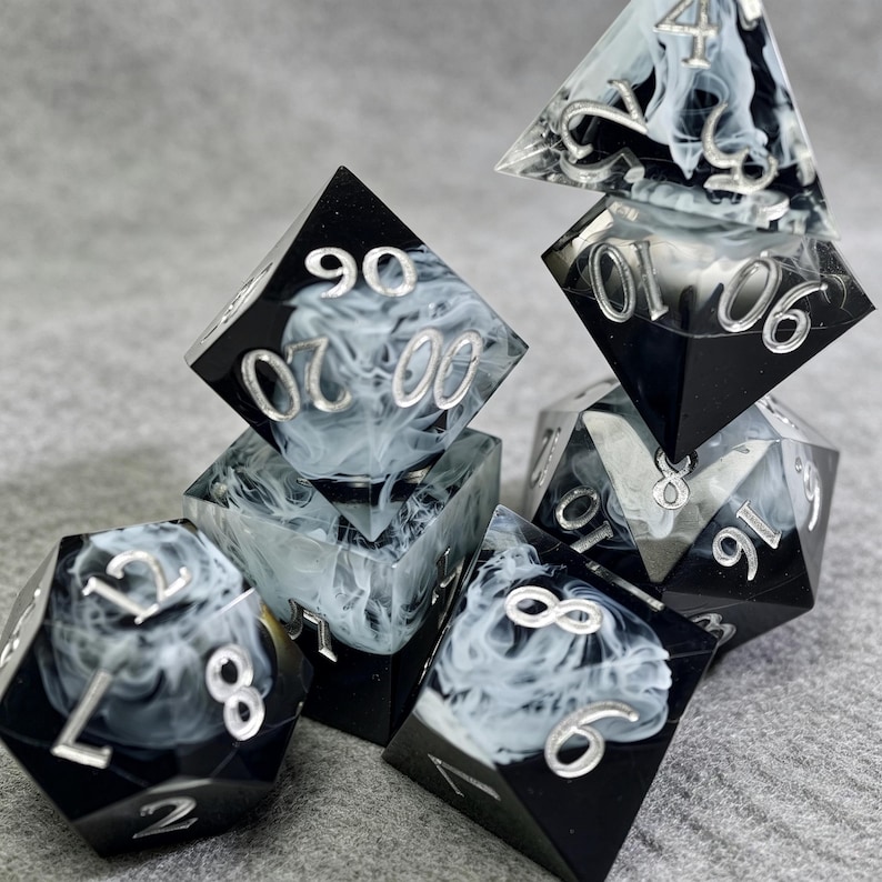 May include: A set of seven polyhedral dice with a black and white smoky design. The dice have silver numbering and are designed for tabletop gaming. The set includes a D4, D6, D8, D10, D%, D12, and D20.