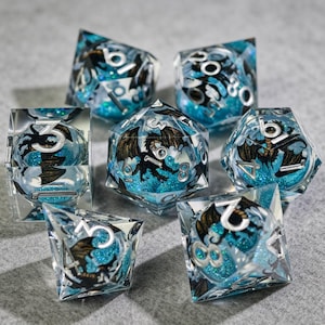 Op de afbeelding: Set van zeven heldere harsdobbelstenen met een drakenontwerp en blauwe glitter. De dobbelstenen hebben zilveren cijfers en een zwarte drakenillustratie. Voor tabletop games.