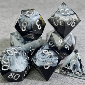 Könnte beinhalten: Ein Satz von sieben Würfeln für Tabletop-Spiele. Die Würfel sind schwarz mit einem wirbelnden weißen Muster und silbernen Zahlen. Das Set enthält einen D4, D6, D8, D10, D12, D20 und einen Prozentwürfel.