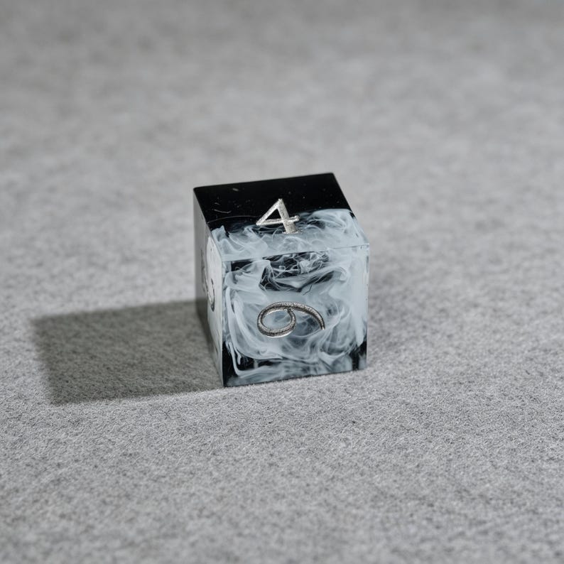 May include: A black and white marbled resin die with silver numbering. The die shows the number 4 on top and the number 6 on the side. The die is a cube shape and is set against a gray background.
