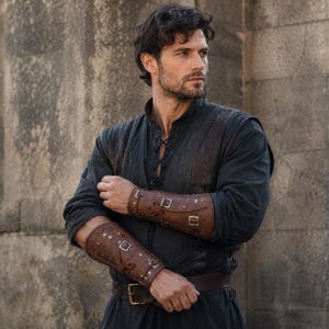 May include: A dark charcoal long-sleeved tunic with a lace-up neckline and matching vest overlay, paired with brown leather forearm bracers featuring silver-toned buckles and rivets, and a brown leather belt with a rectangular metal buckle.
