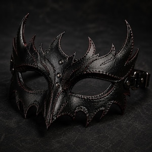 Könnte beinhalten: Eine schwarze Ledermaske mit scharfen, spitzen Kanten und detaillierten dunkelroten Nähten entlang der Ränder. Die Maske verfügt über drei kleine Metallnieten auf dem Nasenrücken sowie einen verstellbaren Riemen mit Metallschnalle an der Seite.