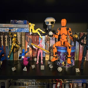 A group of Dummy 13 articulated figures enjoying a session of Dungeons and Dragons with a back drop of Dragonlance novels and Funko Pop dolls of Stan Lee and Warduke. The pink figure holds a pencil, while the orange Titan figure gets ready to roll a d20. A multicolored figure rides on a 3d printed "Benchy."